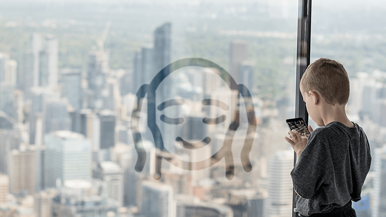 A young white boy stands on the CN Tower's Main Observation Level, against the floor-toceiling window walls, facing out towards the city, holding a smart phone. There's an overlay of the Low Sensory Mornings icon on the picture.