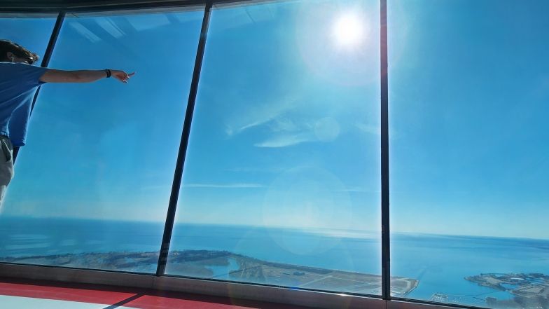 Une personne se tient au niveau d'observation principal de la Tour CN et, à travers les fenêtres allant du sol au plafond, pointe vers le soleil.