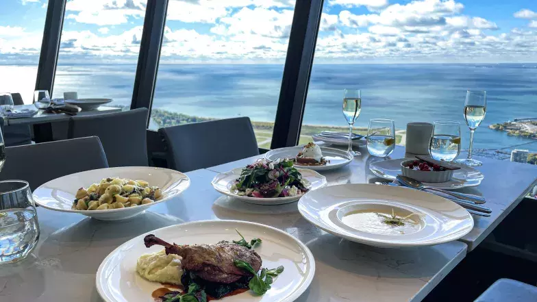 View of multiple dishes on a white marble table at 360 with glasses of water and bubbly on the table as well. All the dishes are part of the Lunch in the Sky menu. In the background, a view of Lake Ontario and Billy Bishop Airport through the floor to ceiling windows.