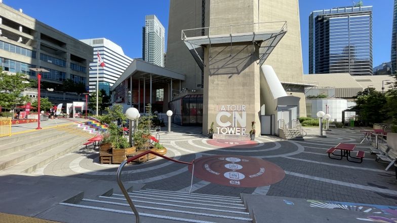 View of the lower patio at the base of the CN Tower. The CN Tower logo sign is in the middle surrounded by plants and flowers. We also see more plants, art and picnic tables all over the lower patio.