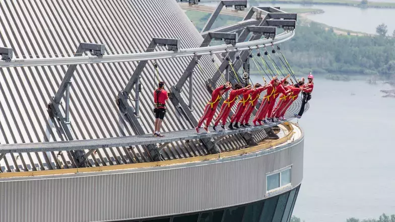 A group of people on the outside of the tower (edgewalk). They are wearing harnesses connected to security lines as they lean forward over the edge of the tower with arms straight out.