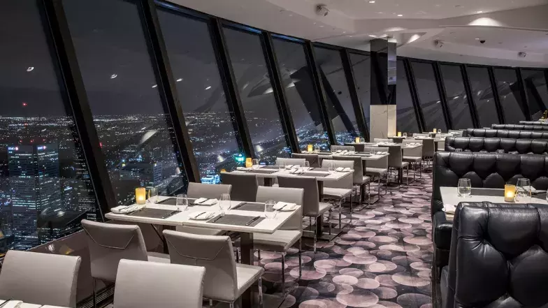 The restaurant window shows Toronto at night. Along the window are tables, chairs. On the other side of the isle are booths. On the tables are lit candle lights. There are no people in the space.