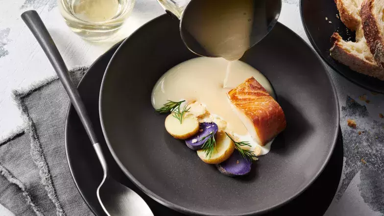 Fish on plate with potato, sauce is being poured on the plate next to the fish and vegetables. A plate of bred and a spoon is on the side of the plate.