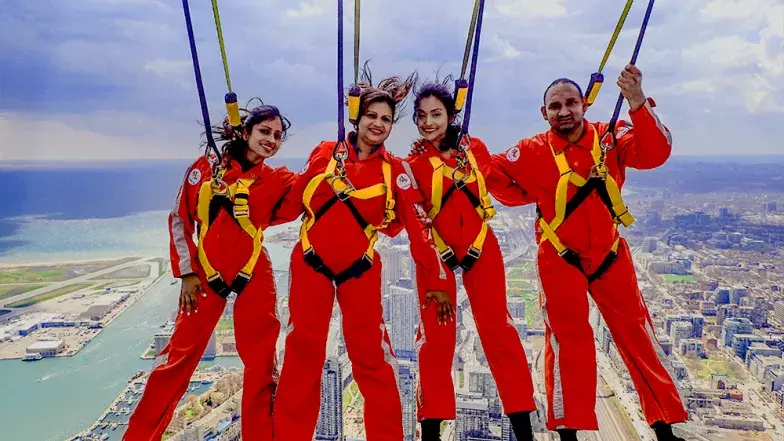 Four people, side by side leaning back over the edge. Far below and behind them is the city of Toronto on a clear summer day