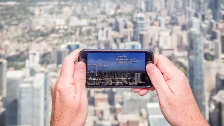 Two hands holding a mobile phone pointed at the City of Toronto from the tower