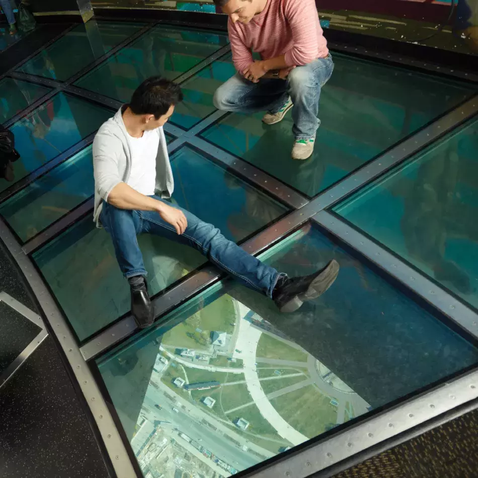 Two people sitting on the glass floor. The ground is visible far below.