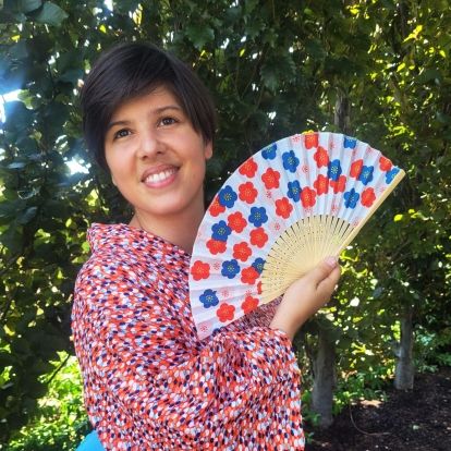 Artist Morgan Kagesheongai who is an Anishinaabe Ojibwe, holding a colourful fan wearing a red and pink floral shirt with trees in the background