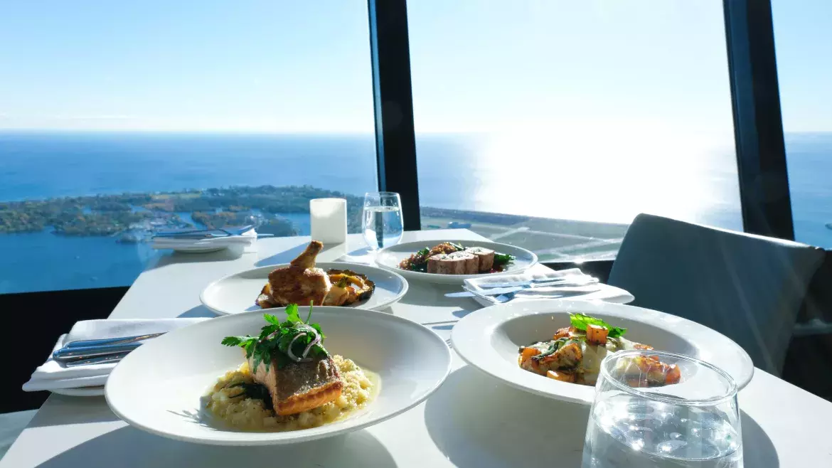 Four dishes from the Winterlicious menu dispersed on a marble table at 360 Restaurant during the day. There are water glasses and cutlery on the table as well. We see Lake Ontario and the Toronto Islands in the background through the floor to ceiling windows.