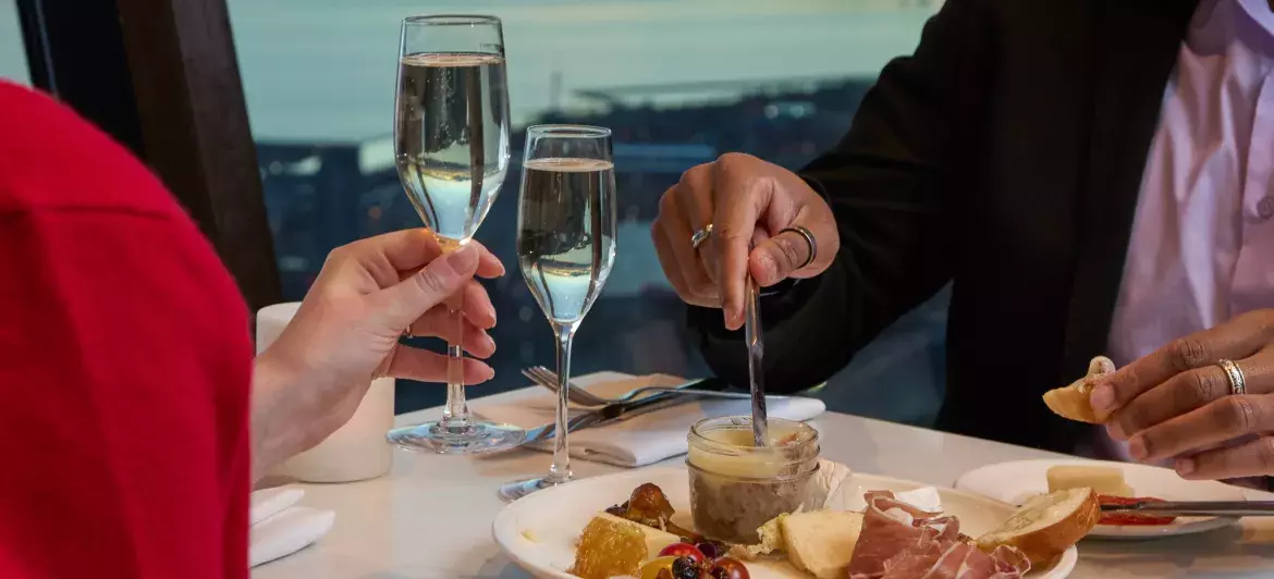 A couple joyfully toasting with champagne at a beautifully set table adorned with a charcuterie board. In the background, we can see the view of Toronto at sunset through the windows.