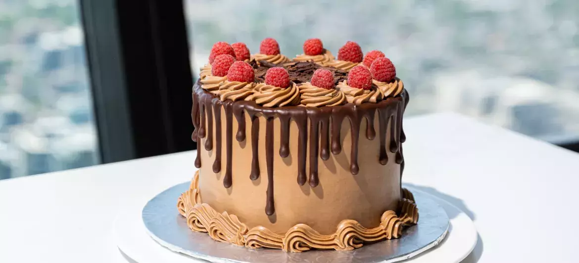 View of a big chocolate cake decorated with raspberries placed on a white marble table at 360 Restaurant. In the background, we can see the view of Toronto through the windows.