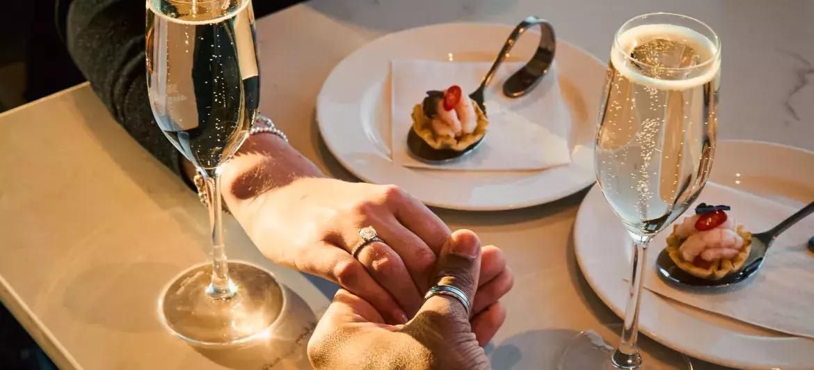 Two hands intertwined over champagne glasses, representing a romantic moment shared between a couple.