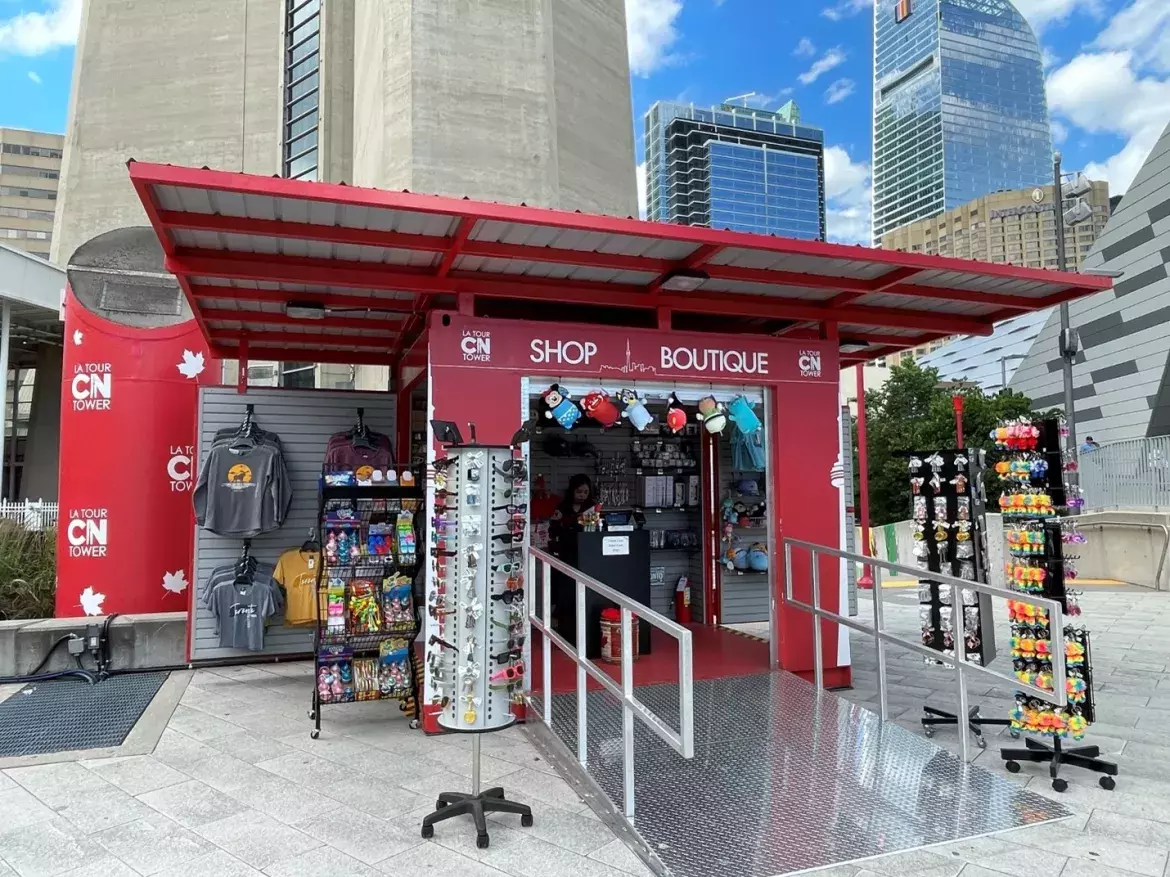 Red outdoor kiosk located in Canada Lands Square with the CN Tower logo and the words Shop/Boutique on the overhead arch. There’s various merchandise like t-shirts, keychains and sunglasses displayed on stands in and around the kiosk.